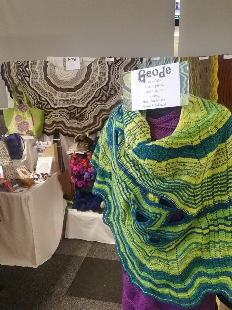 a yarn stand at a show with a tailor's dummy in the foreground draped in a hand-knitted shawl in shades of green 