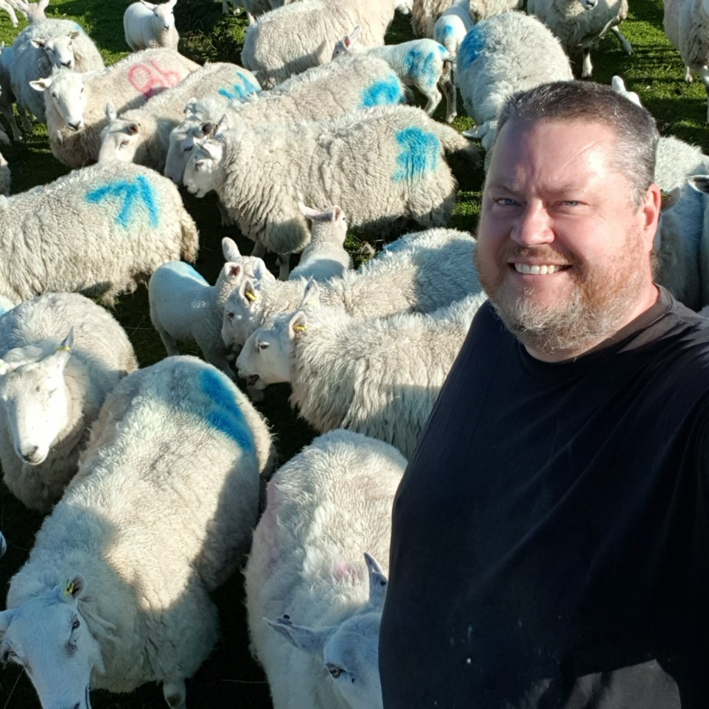 a man stands smiling in front of a group of sheep 