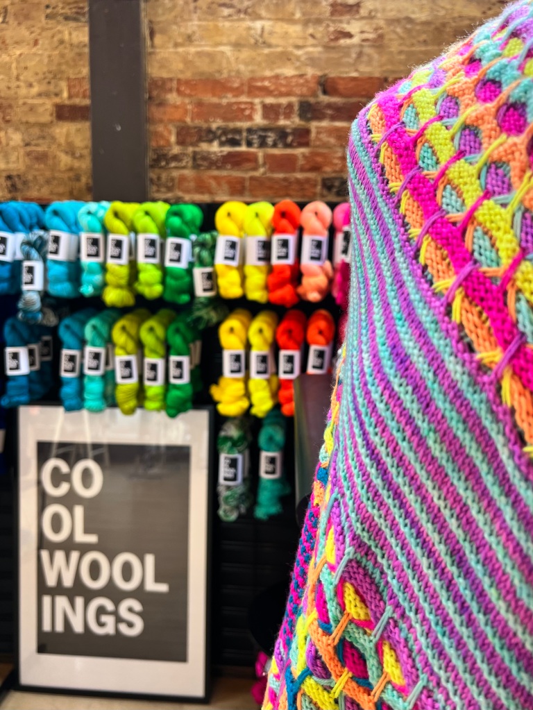 a stand at a yarn show filled with skeins of yarn in bright colours and in the foreground the edge of a knitted shawl in neon shades