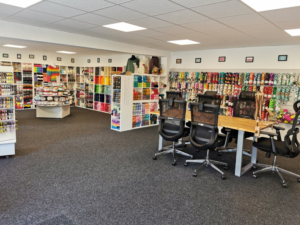 a photo of the inside of the Woolfoll shop, showing a wooden table and chairs in front of a wall of handdyed yarn, in the back corner is cube shelves filled with commercial yarn 