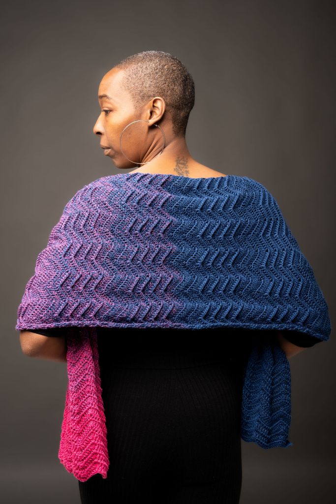 a short-haired black woman stands with her back to the camera and looking off to the side, wearing a crocheted shawl with a chevron pattern