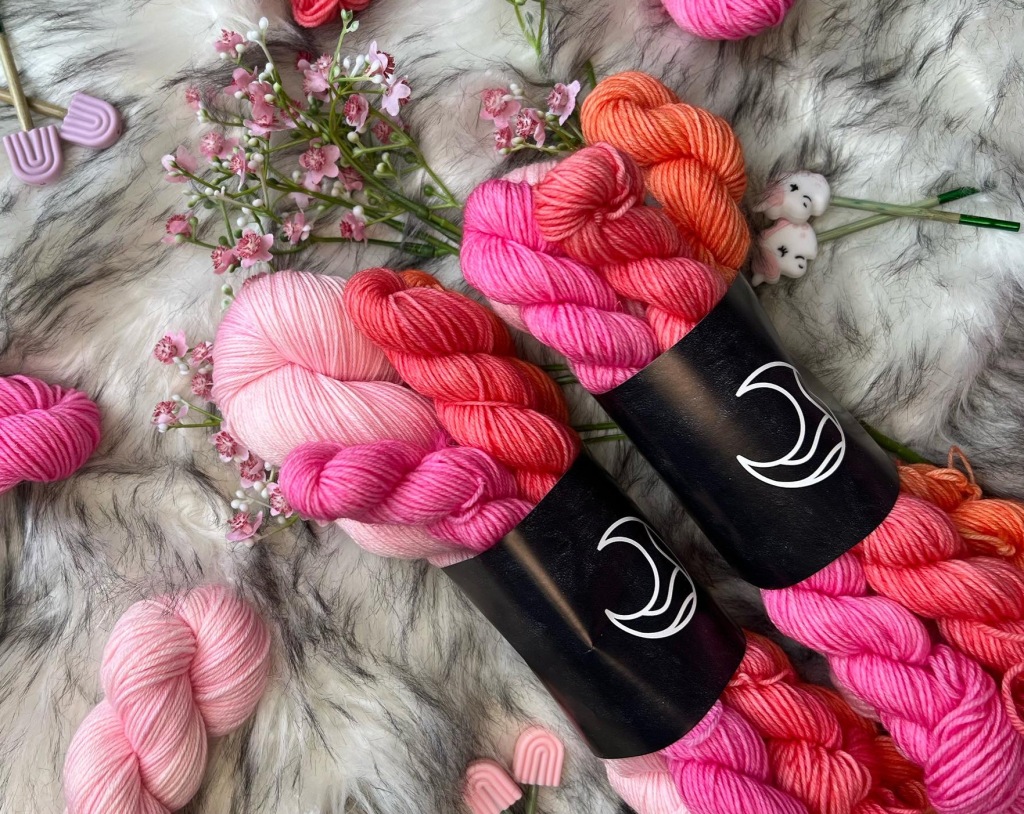 two sock sets of yarn in shades of pink on top of a piece of furry fabric, surrounded by flowers and notions