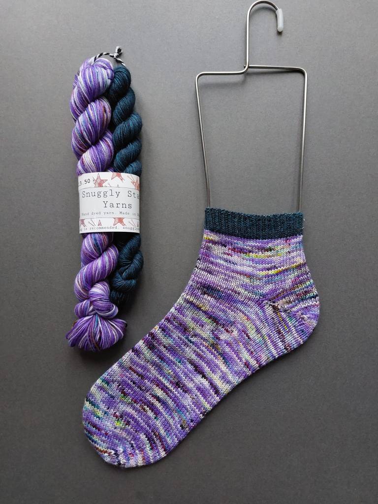 a knitted trainer sock in shades of purple on a metal sock blocker, with the matching yarn alongside