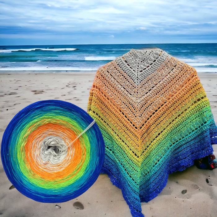 a crocheted shawl in shades of blue, yellow and orange in front of a picture of a beach 
